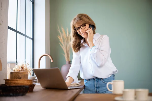 快乐的中年妇女站在厨房的家 有魅力的女性使用笔记本电脑和有一个电话 总部办公室 — 图库照片