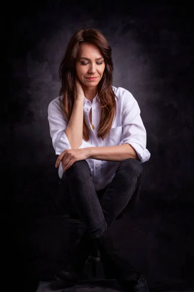 迷人的中年女性 穿着白色衬衫和黑色牛仔裤 靠着孤立的深色背景坐着 复制空间 工作室拍摄 — 图库照片