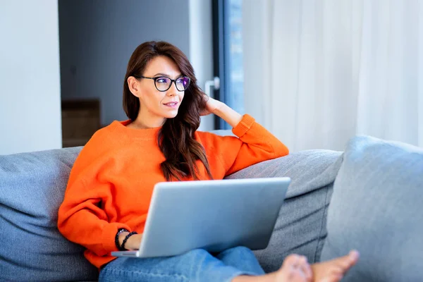 中年妇女坐在沙发上 在网上浏览时 用笔记本电脑射击 自信的女人若有所思地看着 她穿着橙色毛衣和蓝色牛仔裤 — 图库照片