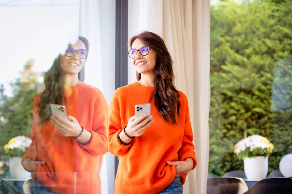 快乐的黑头发女人站在玻璃窗前发短信 穿着橙色毛衣和眼镜的迷人女性 — 图库照片