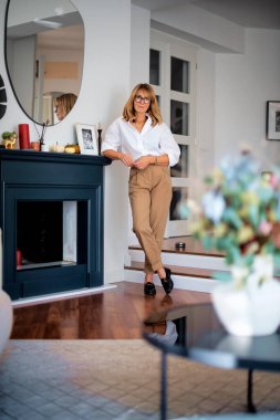 Beyaz gömlek ve pantolon giyen, şöminenin yanında dikilen çekici, sarışın bir kadın..