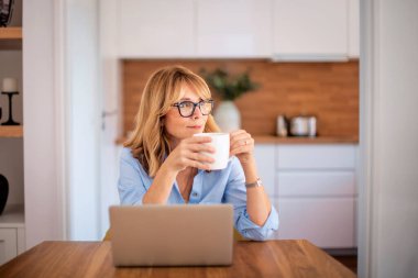 Mutfakta masada otururken çay içen kendinden emin bir kadın. İş için dizüstü bilgisayar kullanan orta yaşlı bir kadın. Merkez Ofis. 