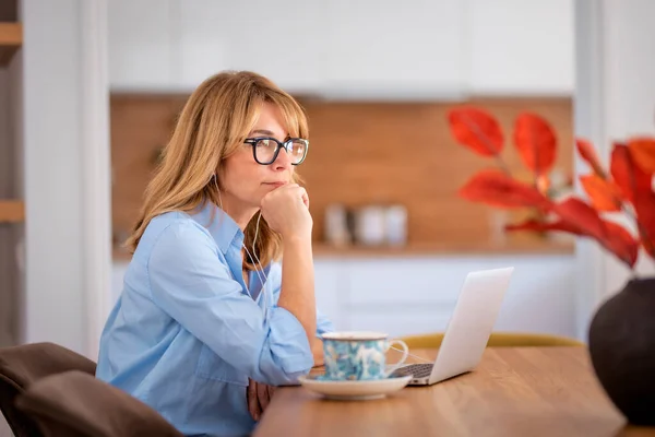 思考中年妇女使用笔记本电脑和有视频通话 同时在家工作 自信的女人坐在厨房的桌旁 总部办公室 — 图库照片