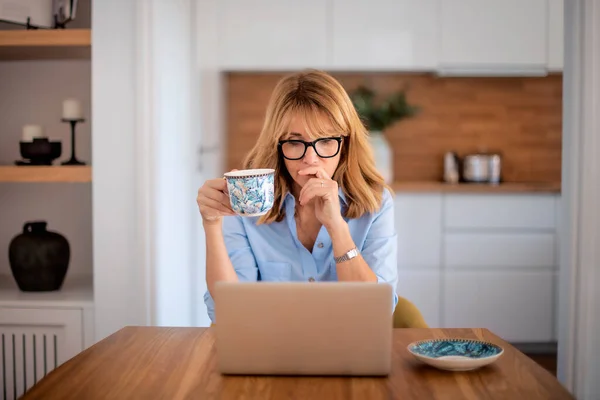 想着中年妇女在家工作的时候用笔记本电脑喝茶 自信的女人坐在厨房的桌旁 总部办公室 — 图库照片