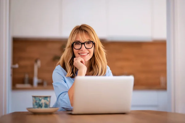 用笔记本电脑微笑中年妇女 在家工作 自信的女人笑着坐在厨房的桌旁 总部办公室 — 图库照片