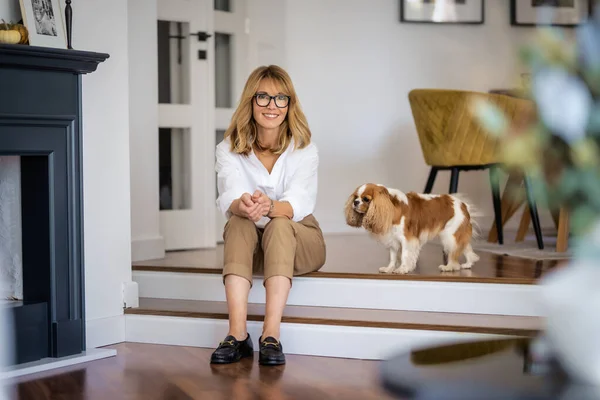 满满的快乐中年女人坐在地板上 白色的可爱的小狗 在家里放松的时候 迷人的女人戴着眼镜 穿着休闲装 — 图库照片