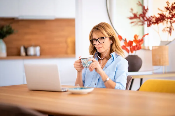 中年の女性は彼女のダイニングルームのテーブルに座って ビデオ通話をしながらイヤホンやノートパソコンを使用しています 内務省 — ストック写真