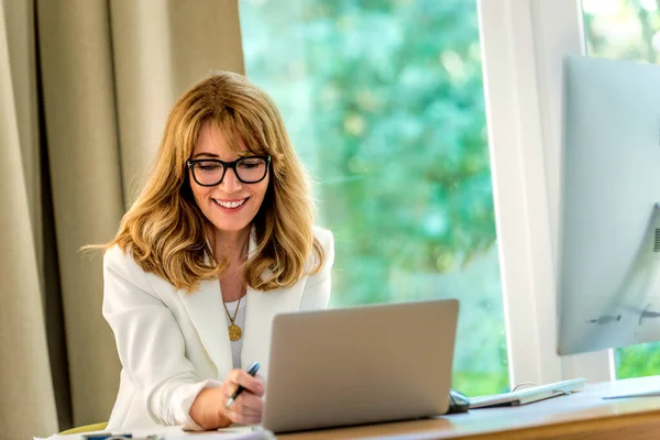 一个快乐的微笑的中年女商人坐在办公桌前 用笔记本电脑工作 自信的职业女性穿着白色夹克和眼罩 总部办公室 — 图库照片