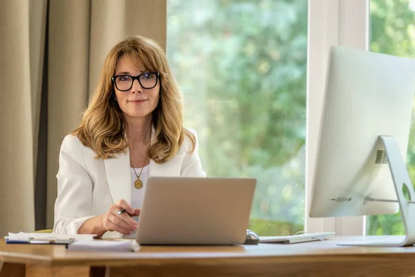微笑着坐在办公桌前 用笔记本电脑工作的中年女商人 自信的职业女性穿着白色夹克和眼罩 总部办公室 — 图库照片