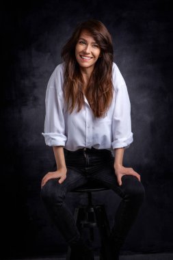 Çekici bir kadının stüdyo portresi. Gömlek giyip gülerken izole koyu gri arka planda oturuyor. Boşluğu kopyala. 