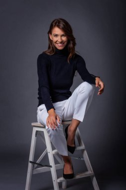 Gri arka planda oturan beyaz dişli, gülümseyen güzel bir kadın. Balıkçı yaka kazak giyen mutlu kadın. Tam yol ileri. Boşluğu kopyala.