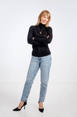 Siyah kazak ve kot pantolon giyen çekici, sarı saçlı bir kadın. Kollarını kavuşturmuş neşeli, gülümseyen bir kadın. Gri arka plana karşı. Boşluğu kopyala.