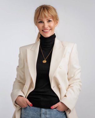 Beyaz ceket ve siyah kazak giyen çekici sarı saçlı bir kadın. Gri arka planda duran gülümseyen kadın. Boşluğu kopyala.