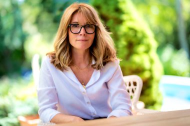 Evdeki terasta dinlenirken orta yaşlı, gülümseyen, gözlüklü ve beyaz gömlekli bir kadının yakın plan portresi. Çekici bir kadın, sarı saçlı ve kameraya bakıyor.. 