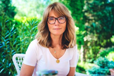 Evdeki terasta dinlenirken orta yaşlı, gülümseyen, gözlüklü ve beyaz gömlekli bir kadının yakın plan portresi. Çekici bir kadın, sarı saçlı ve kameraya bakıyor.. 