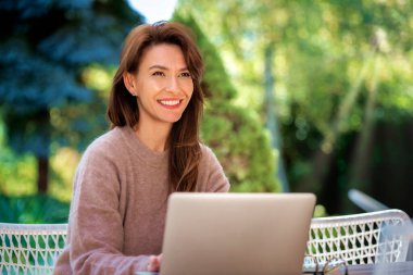 Neşeli, gülümseyen esmer saçlı bir kadın terasta dizüstü bilgisayar kullanıyor. Bilgisayarda çalışan çekici bir kadın ya da görüntülü arama. Merkez Ofis.