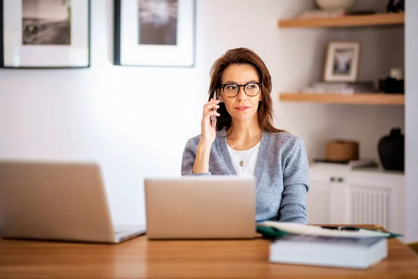 迷人的黑发女人坐在家里 用手提电脑打电话 自信的女性穿着休闲装和眼镜 回家去吧 免版税图库图片