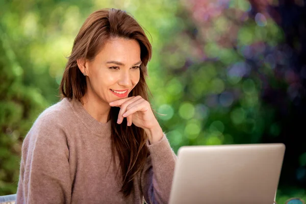 一位快乐的 面带微笑的黑发女人正坐在阳台上使用笔记本电脑 迷人的女性在笔记本电脑上工作或有视频通话 总部办公室 图库照片