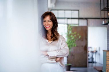 Kahverengi saçlı, orta yaşlı, neşeli bir kadının portresi. Çekici bir kadın kameraya bakıyor ve kolları pencerede çapraz duruyor.. 