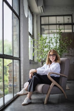 Pencere kenarındaki koltukta oturan çekici bir kadının boyu. Beyaz gömlekli, siyah pantolonlu ve neşeli gülümseyen orta yaşlı bir kadın.. 