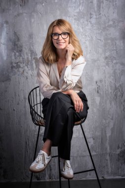 Gri bir duvarın önünde oturan güzel sarı saçlı bir kadın. Gömlek ve pantolon giyen ve kameraya bakan gülümseyen bir kadın. Tam yol ileri. Boşluğu kopyala.