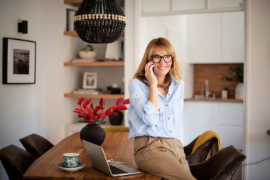 Orta yaşlı bir kadın yemek masasında oturuyor ve telefonla konuşuyor. Gündelik kıyafetler giyen ve neşeli gülümseyen çekici bir kadın. Merkez Ofis. 