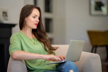 Esmer bir kadın evdeki koltukta oturuyor ve internette video konferans veya geziniyor. Genç bir bayan günlük kıyafetler giyiyor ve gülümsüyor. Merkez Ofis.