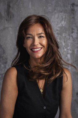 Gri bir duvarın önünde duran güzel kahverengi saçlı bir kadının yakın çekimi. Gülümseyen, siyah tişörtlü ve başka tarafa bakan neşeli bir kadın. Saçını üflüyordu. Boşluğu kopyala.