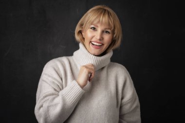 Siyah arka planda duran orta yaşlı bir kadın. Kendinden emin kadın balıkçı yaka kazak giyiyor. Sarı saçları var..