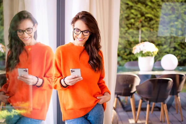 迷人的中年妇女正站在她家的窗前 她穿着橙色的毛衣和眼镜 用的是智能手机 — 图库照片