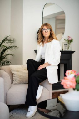 Orta yaşlı, çekici bir kadın evdeki koltukta dinleniyor. Sarı saçlı, gözlüklü ve beyaz ceketli bir kadın. Tam boy çekim. 