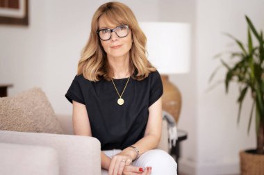 Orta yaşlı, çekici bir kadın evdeki koltukta oturuyor. Sarı saçlı, gözlüklü, sıradan kıyafetler giyen bir kadın..