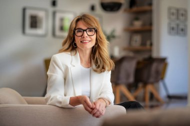 Orta yaşlı, çekici bir kadın evdeki koltukta dinleniyor. Sarı saçlı, gözlüklü ve beyaz ceketli bir kadın..