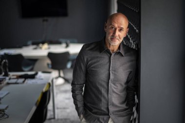 Ofiste duran orta yaşlı bir işadamı duvara yaslanıyor. Gömlek giyen ve kameraya bakan profesyonel bir adam..