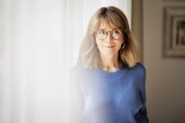 Pencerenin önünde duran orta yaşlı güzel bir kadının portresi. Sarı saçlı, mavi gömlekli, gözlüklü ve kameraya bakan bir kadın. Boşluğu kopyala.