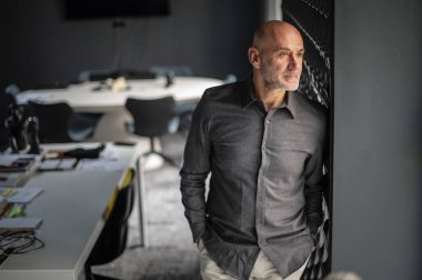 Ofiste duran orta yaşlı bir işadamı duvara yaslanıyor. Tişört giyen ve düşünceli görünen profesyonel bir adam..