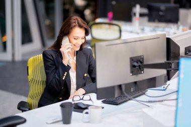 Orta yaşlı iş kadını bilgisayar kullanıyor ve ofiste masa başında otururken telefonla konuşuyor. Finansal raporlar üzerinde çalışan profesyonel bir kadın..