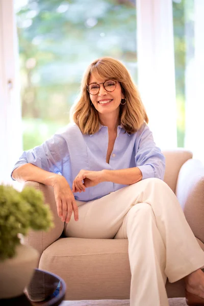 一个金发美女的画像正坐在家里的扶手椅上 笑着穿蓝衬衫和白裤子的女人 — 图库照片