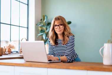 Sabahları mutfak tezgahında dizüstü bilgisayar kullanan orta yaşlı bir kadın. Kendine güveni tam bir kadın günlük kıyafetler giyiyor ve video görüşmesi yapıyor. Merkez Ofis.