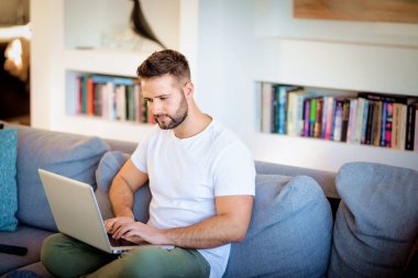 Evdeki kanepede oturan kendine güvenen adam iş için dizüstü bilgisayar kullanıyor. Gündelik kıyafetler giyen yakışıklı bir erkek. Merkez Ofis. 