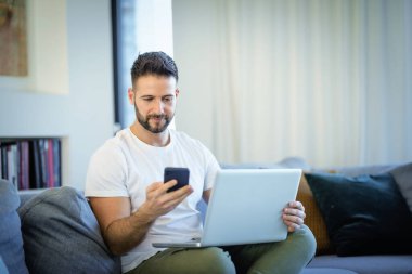 Evde oturan yakışıklı adam dizüstü bilgisayar ve cep telefonu kullanıyor. Orta yaşlı erkek günlük kıyafetler giyiyor ve evde çalışıyor. Merkez Ofis. 