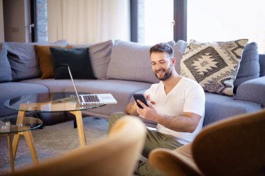 Evde oturan yakışıklı adam dizüstü bilgisayar ve cep telefonu kullanıyor. Orta yaşlı erkek günlük kıyafetler giyiyor ve evde çalışıyor. Merkez Ofis. 