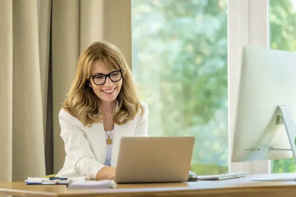 ブロンド髪の実業家が机の上に座ってノートパソコンに取り組んでいる プロフェッショナルな女性がアイウェアとホワイトブレーザーを着用しています ホームオフィス ストック画像