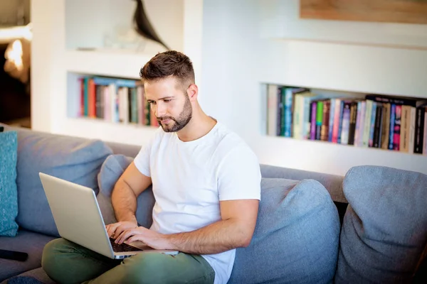 自信的男人坐在家里的沙发上 用笔记本电脑工作 英俊的男性穿着休闲装 总部办公室 免版税图库图片
