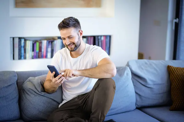 微笑的男人放松在沙发上的家和使用智能手机 穿着休闲装和发短信的自信男性 免版税图库图片