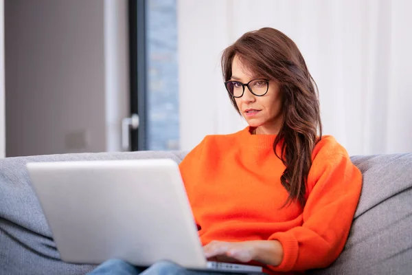 真剣な顔のブルネットハイレッド女性は 自宅でソファーに座って 仕事のためにラップトップを使用しています 魅力的な女性はオレンジ色のセーターを着ています ホームオフィス ロイヤリティフリーのストック写真
