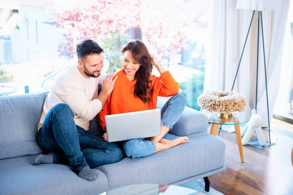 若いカップルが自宅のソファーでノートパソコンを使っている カジュアルな服を着て インターネットでブラウジングしたり ビデオ通話をしたりする幸せな女性と男性 ストック画像