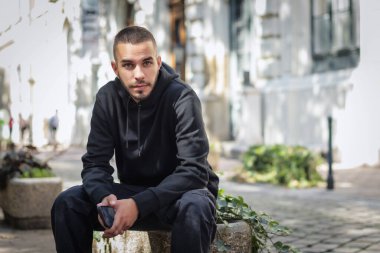 Şehirde sokakta dinlenen yakışıklı bir gencin portresi. Kapüşonlu ve kameraya bakan kendine güvenen bir adam..