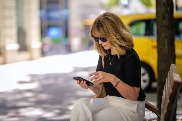 迷人的金发女人坐在街上的长椅上 用手机 戴着太阳镜和发短信的快乐微笑的女性 — 图库照片
