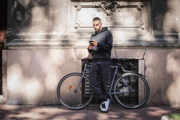 一个英俊的年轻人站在城里的街上用他的手机打了整整一个小时的电话 年轻人骑着自行车在墙边放松 发短信 — 图库照片